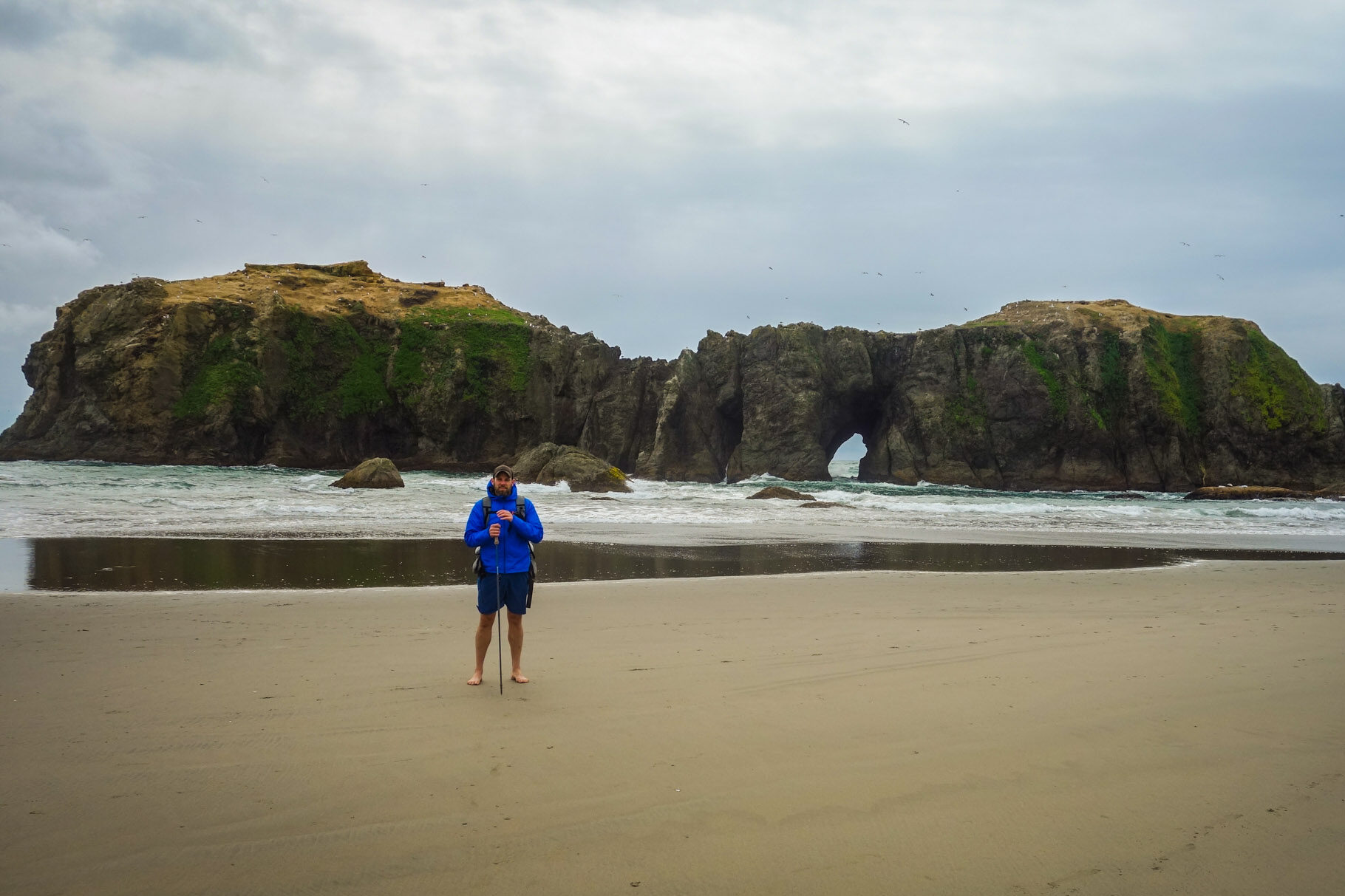 A barefoot backpacker posing in front of a shorline rock formation with a natural bridge on it on the oregon coast trail.