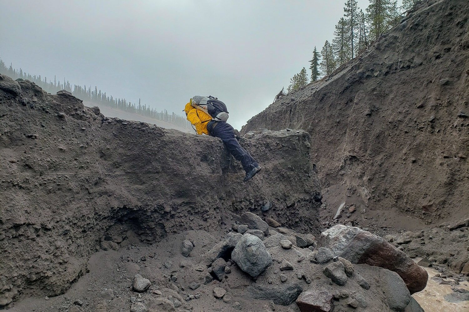 backpacker hoisting themselves up a washed out rugged river bank that is taller than the hiker. 