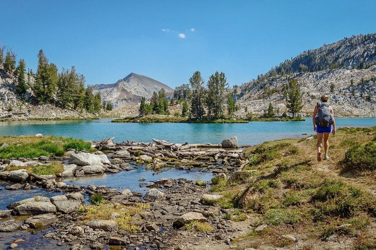 A backpacker hikes by the edge of a turquoise blue lake in the Wallowa mountains with a beautiful island in the middle