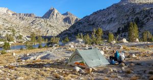 a backpacker seated next to their zpacks triplex tents near an alpine lake in california