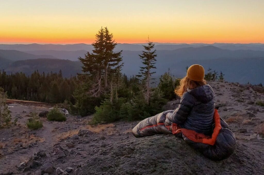 A backpacker sitting on a boulder in the REI Magma 30 sleeping bag while overlooking a mountain view at sunset