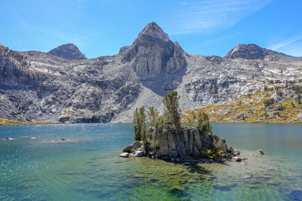 Small island with trees surrounded by clear blue green water sounded by rocky mountains on the John Muir Trail