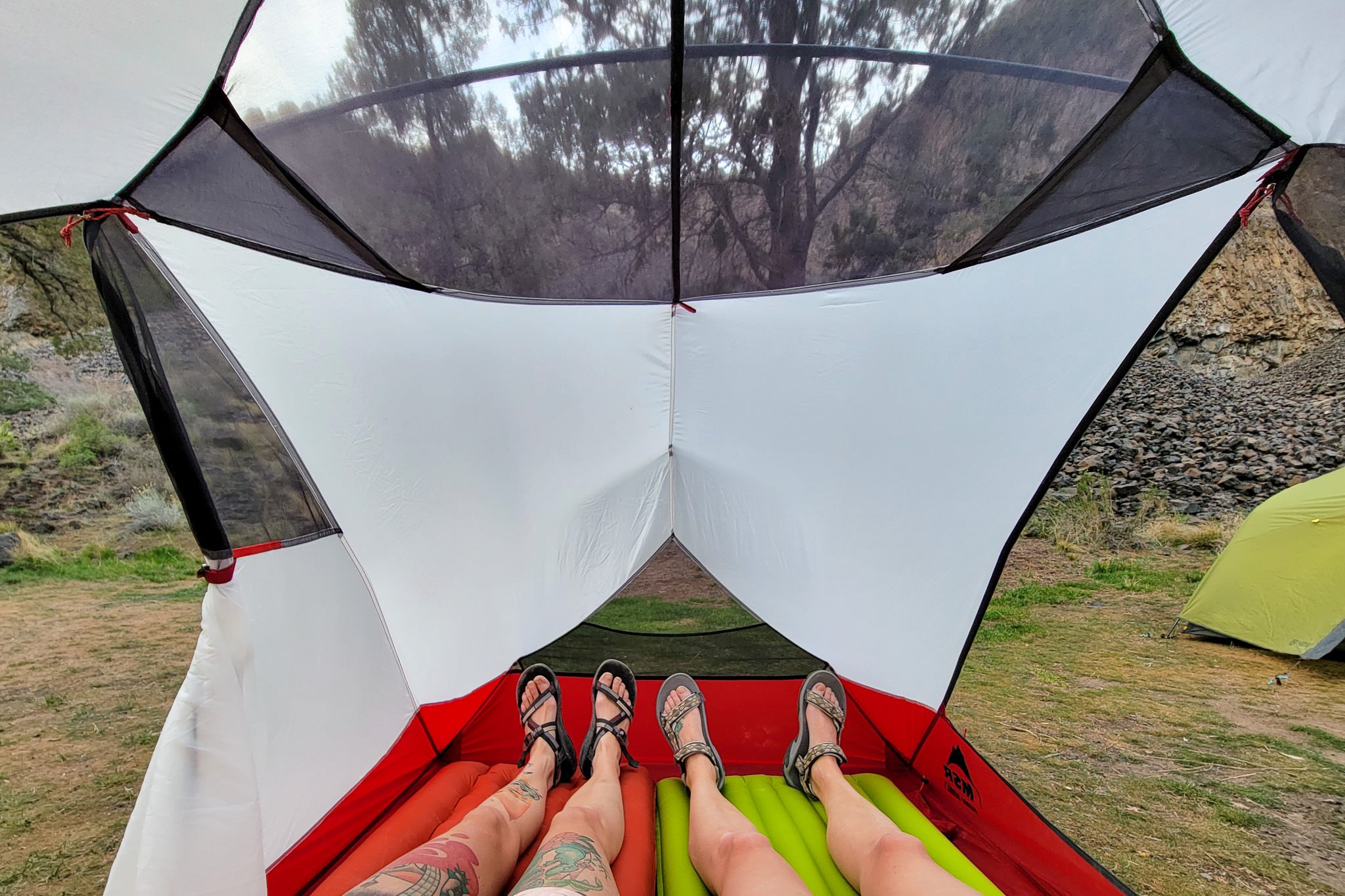 Picture from inside a tent looking towards the feet of two people, the mesh of the inner tent, and the woods outside.