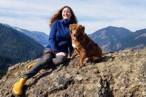 a hiker wearing a fleece jacket while seated on a rocky mountain next to their dog smiling for the camera. Mountains rise in the background.