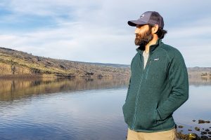 a man with a cap on wearing a patagonia fleece zip jacked in green overlooking a flat body of water