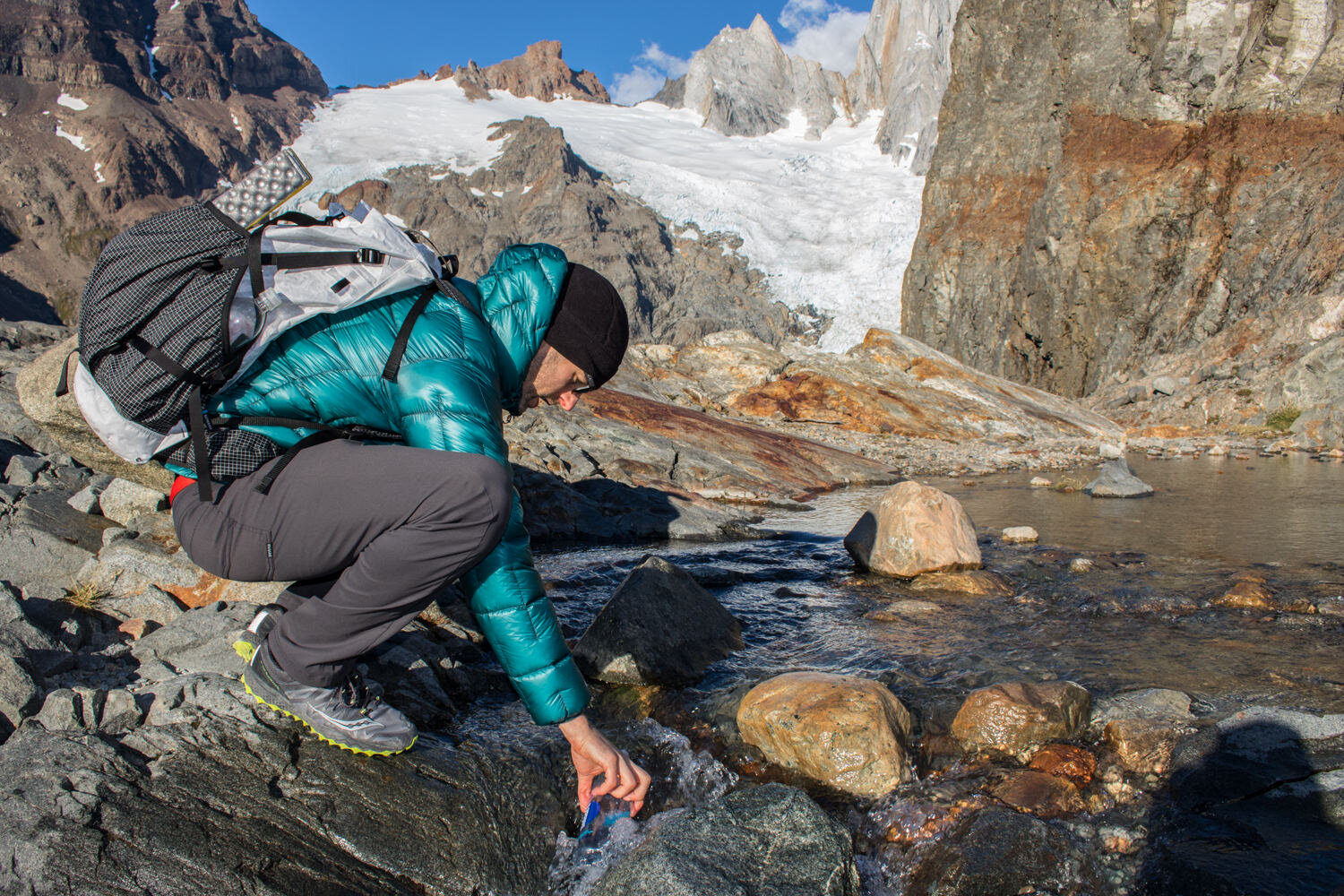 A backpacker bending down to collect water from a glacial stream in the Prana Brion Pants