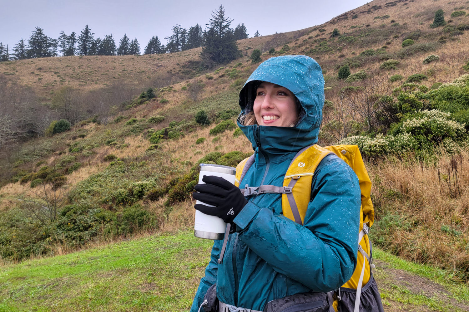 A smiling female hiker in a green Black Diamond Stormline Stretch rain jacket on a coastal hike