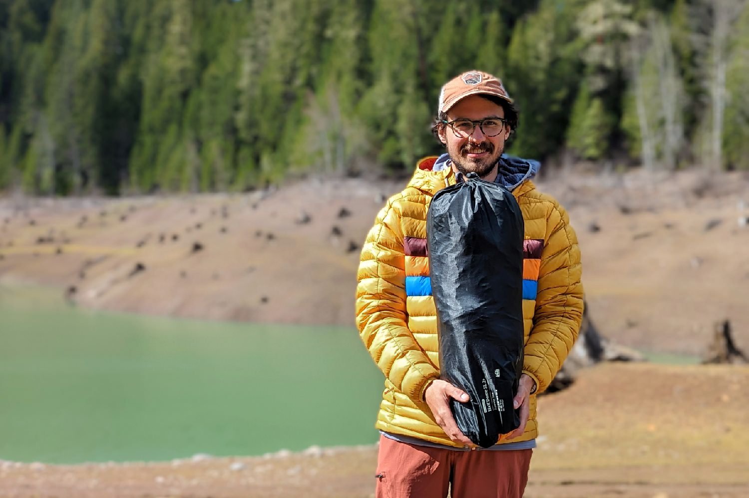 A hiker facing forward and smiling holding the REI Half Dome 2 packed up in its stuff sack - there is a lake and some pine trees in the background