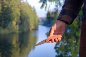 a pocket knife held open in a hiker's hand in front of a bright blue river or lake with evergreens surrounding the banks