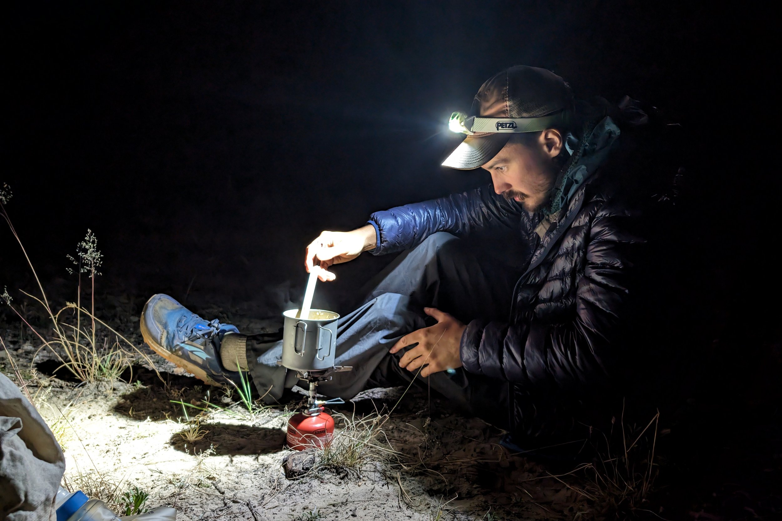 a backpacker making dinner in the dark with their headlamp shining on the camp stove with his spoon stirring the pot