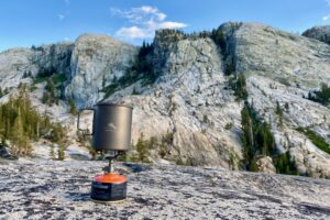 A small backpacking stove boils a pot of water with rugged rocky mountains in the background