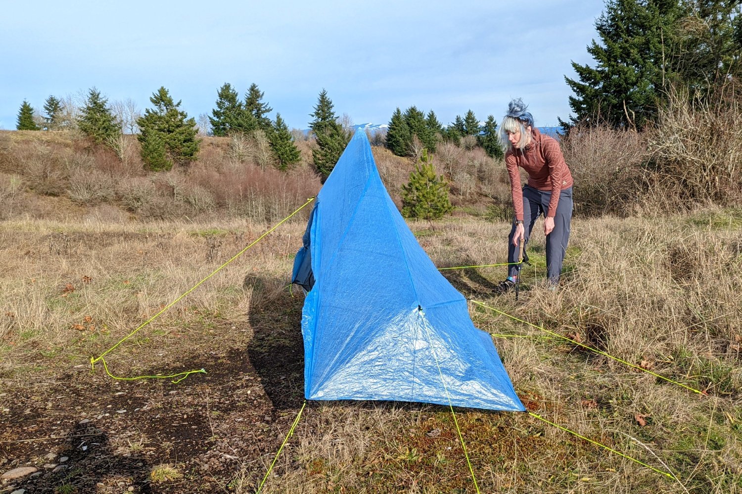 a backpacker pulling and staking guylines on their zpack plex solo tent pitched in a meadow