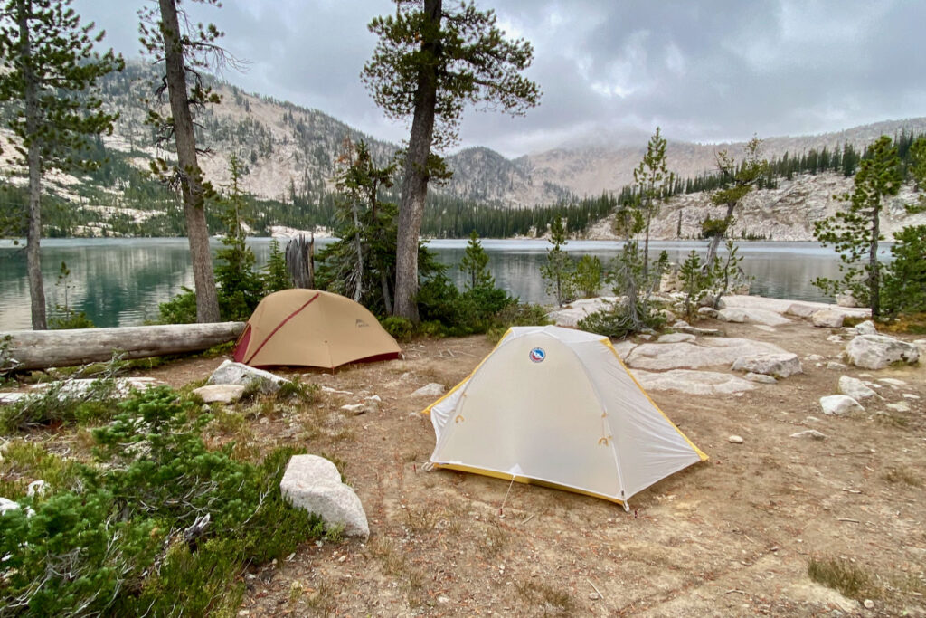 Two tents pitched in a campsite near an alpine lake with mountains in the background.