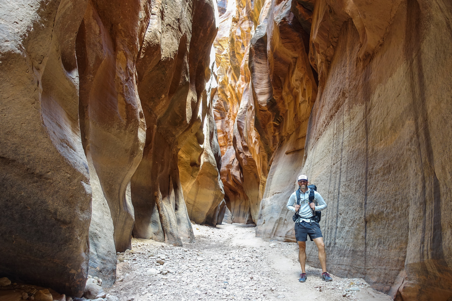 a backpacker stops a poses for the camera below some wavy walls of a slot canyon in utah