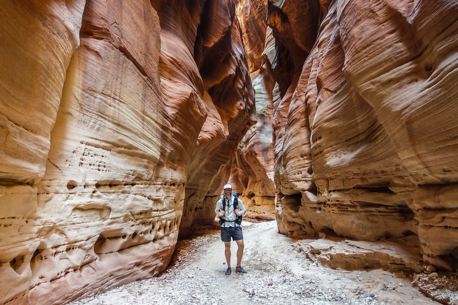 a hiker in paria canyon poses in a slot where the walls have eroded holes and pockets and shelves along the sidewalls.