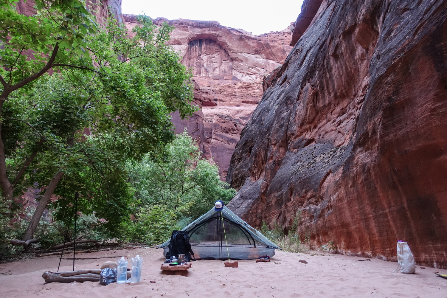 a red sandbar campsite with water containers, packs, and a tent set up with red rocks and green trees in the background