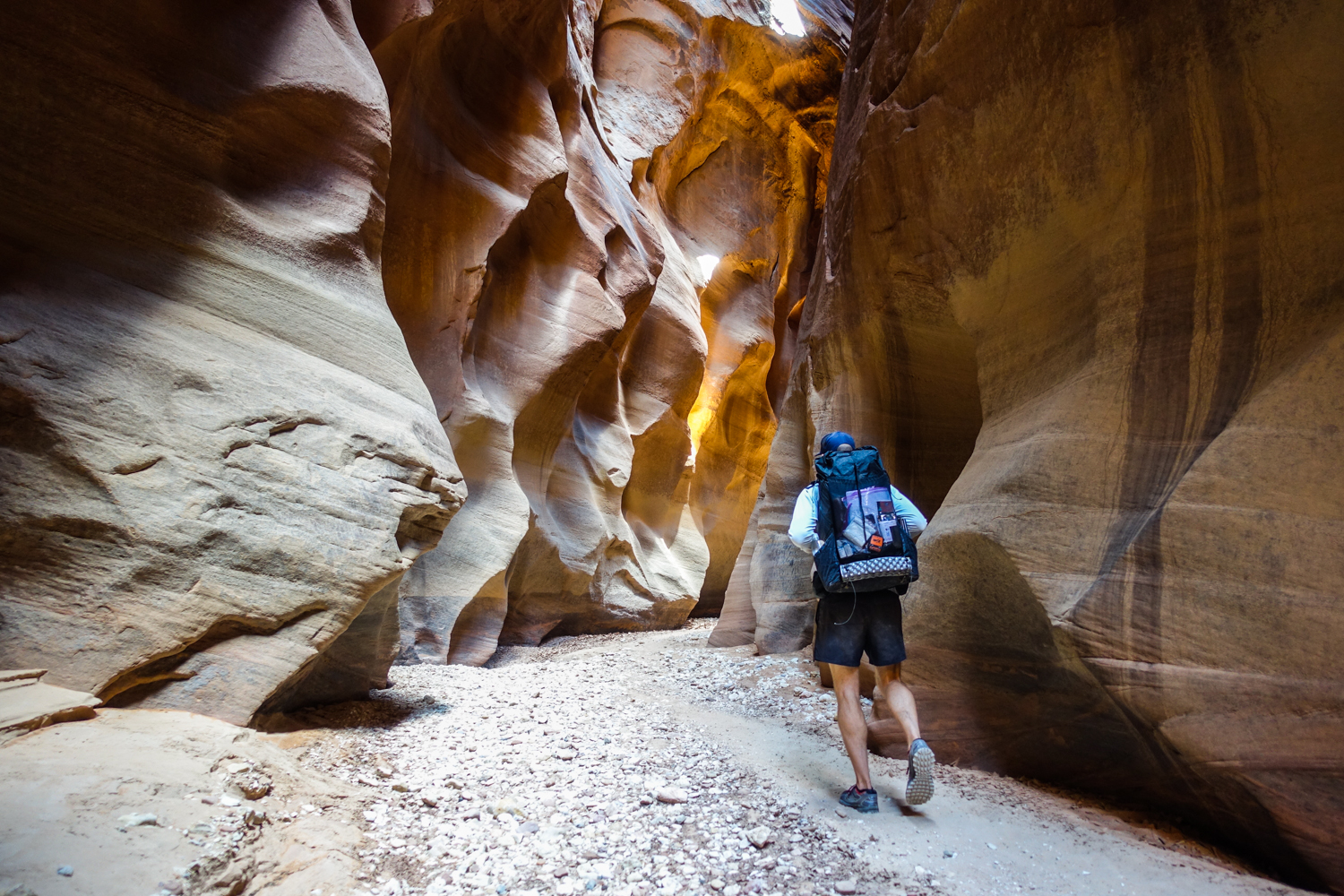 a hiker walking through a slot canyon where the river eroded the walls into wavy red and orange pockets and striations.