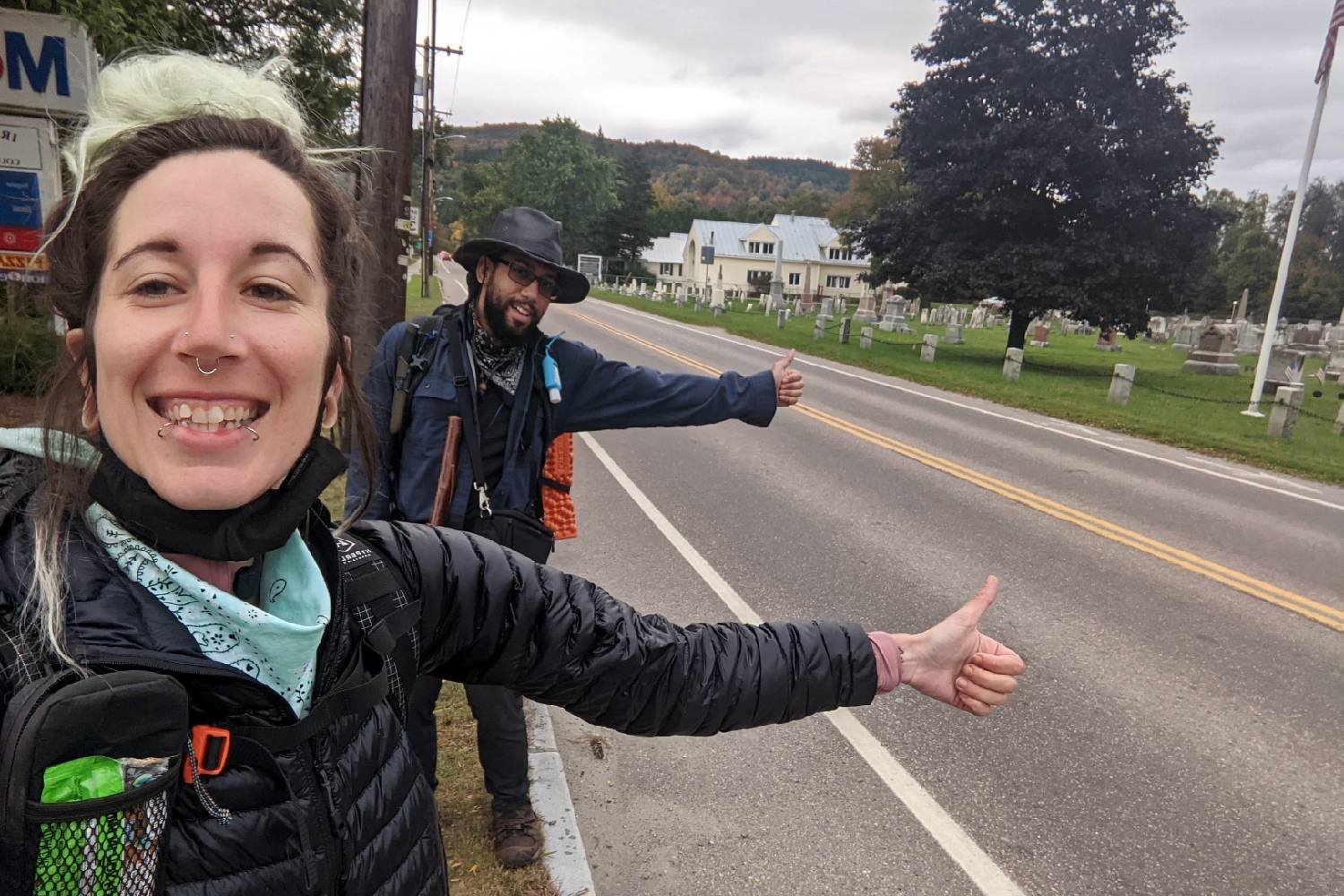 Two Appalachian Trail thru-hikers holding their thumbs out to hitch a ride in a rural town.