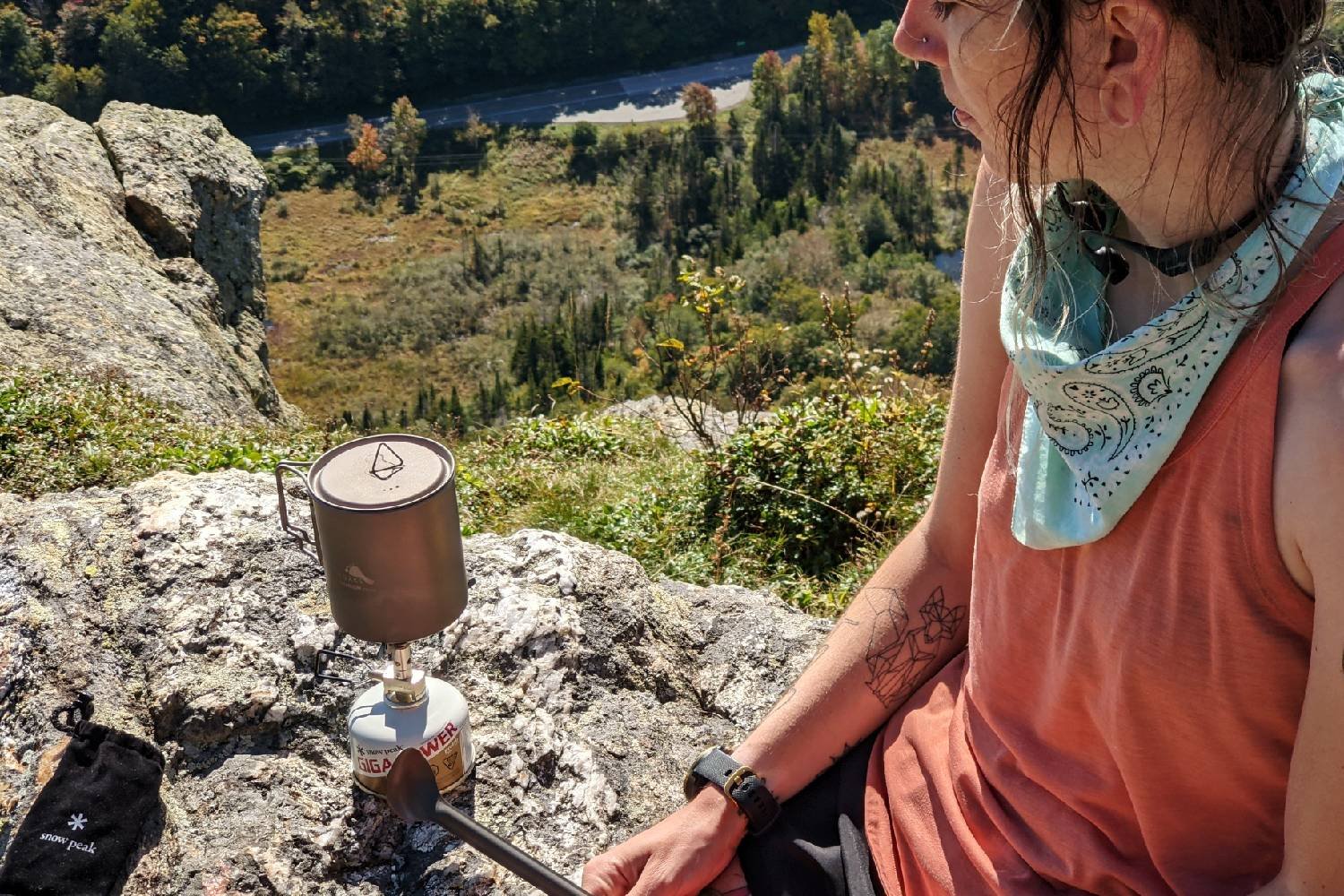 Appalachian Trail thru-hiker making a meal with a camping stove on a ledge overlooking fall foliage and a scenic highway.