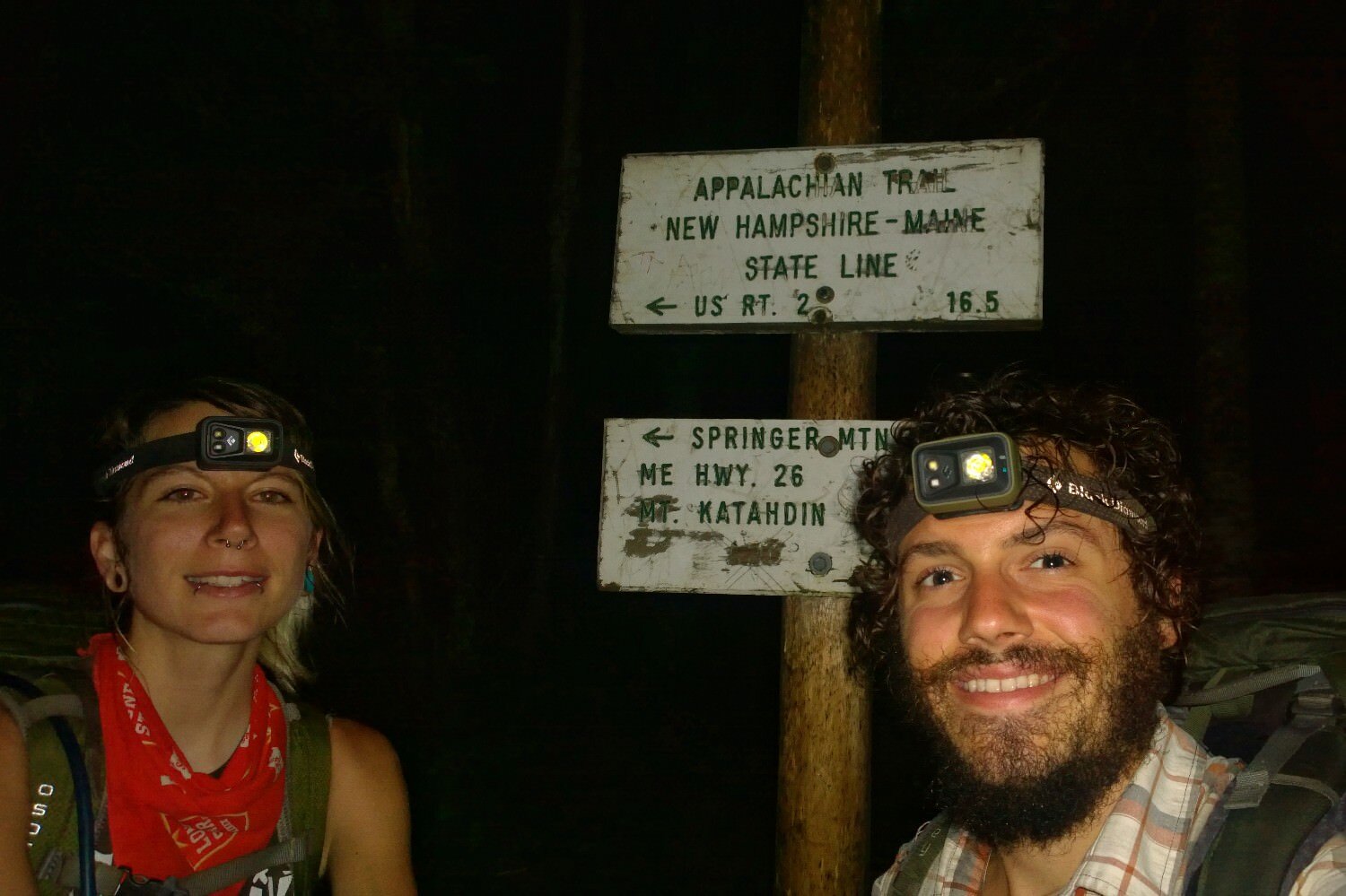 two thru-hikers pose in front of a trail entrance with headlamps on to hike in the dark