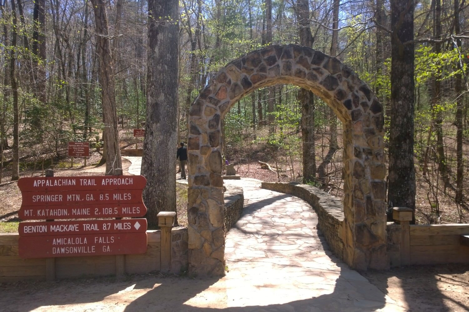 Trailhead entrance with a stone arch and trail signs on a sunny spring day.