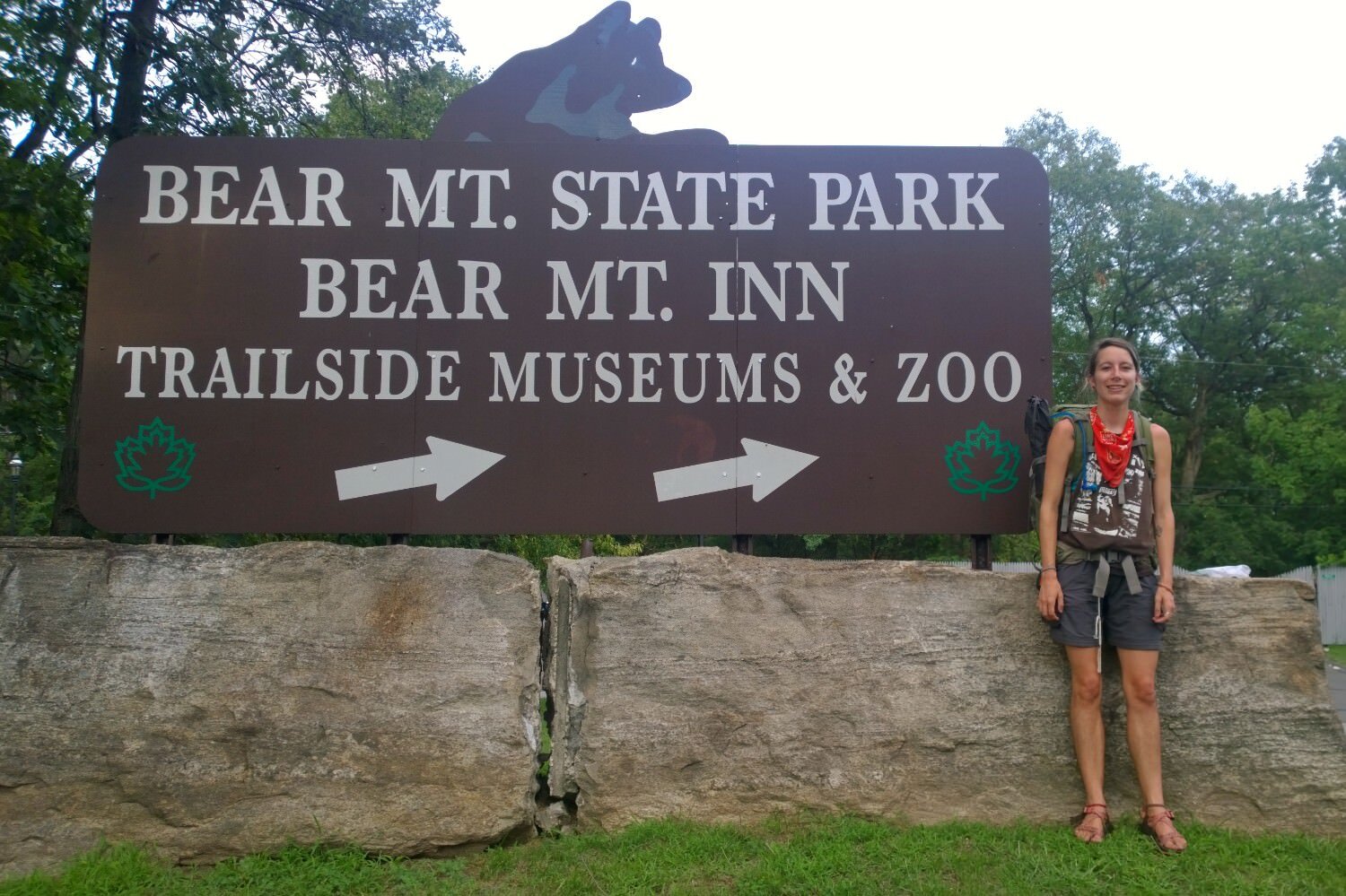 Appalachian Trail thru-hiker posing in front of a large sign stating Bear Mt. State Park, Bear Mt. Inn, trailhead museum & zoo.