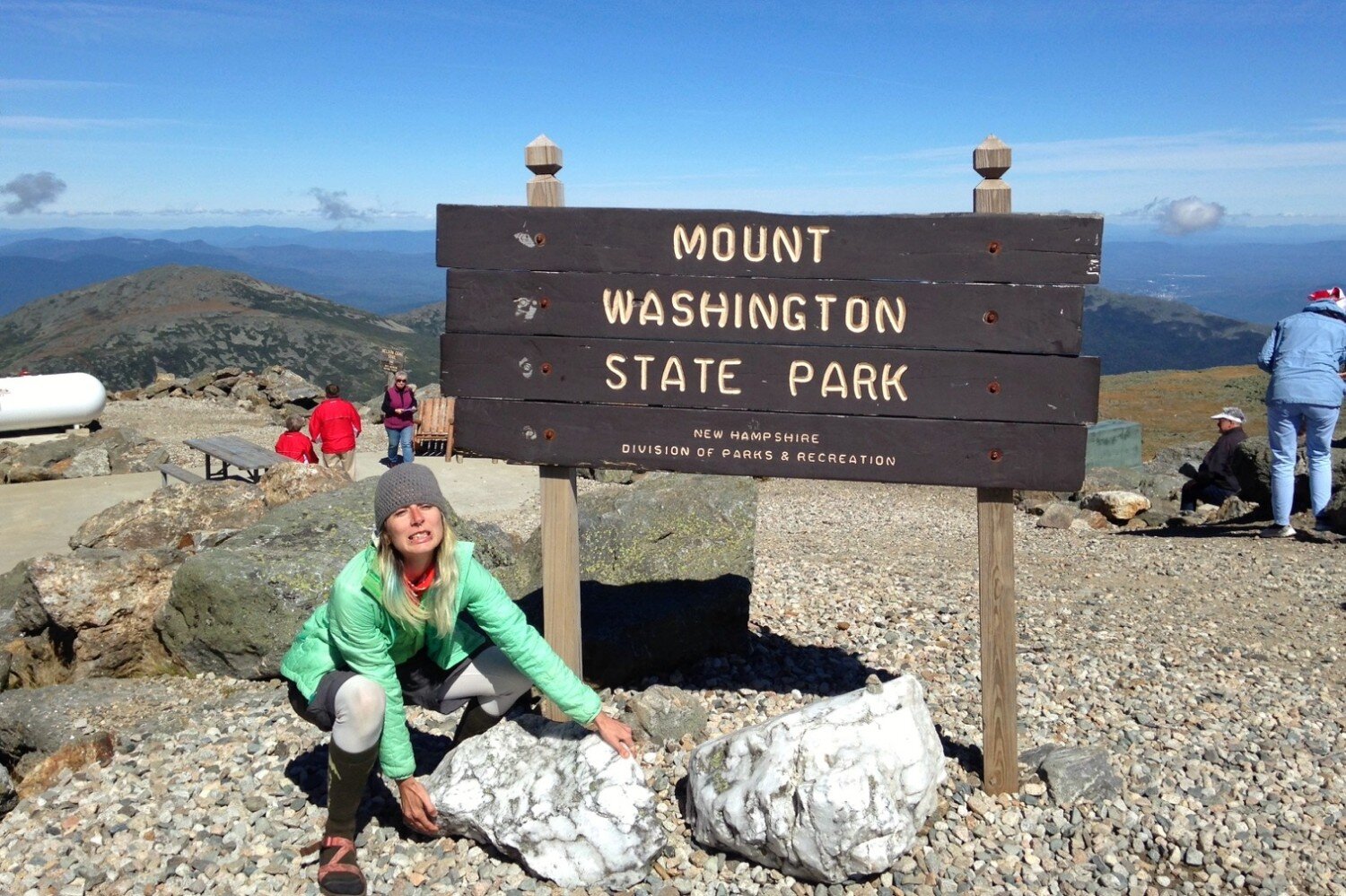 Appalachian Trail thru-hiker pretends to pick up a boulder on top of Mount Washington with a brown sign stating Mount Washington State Park