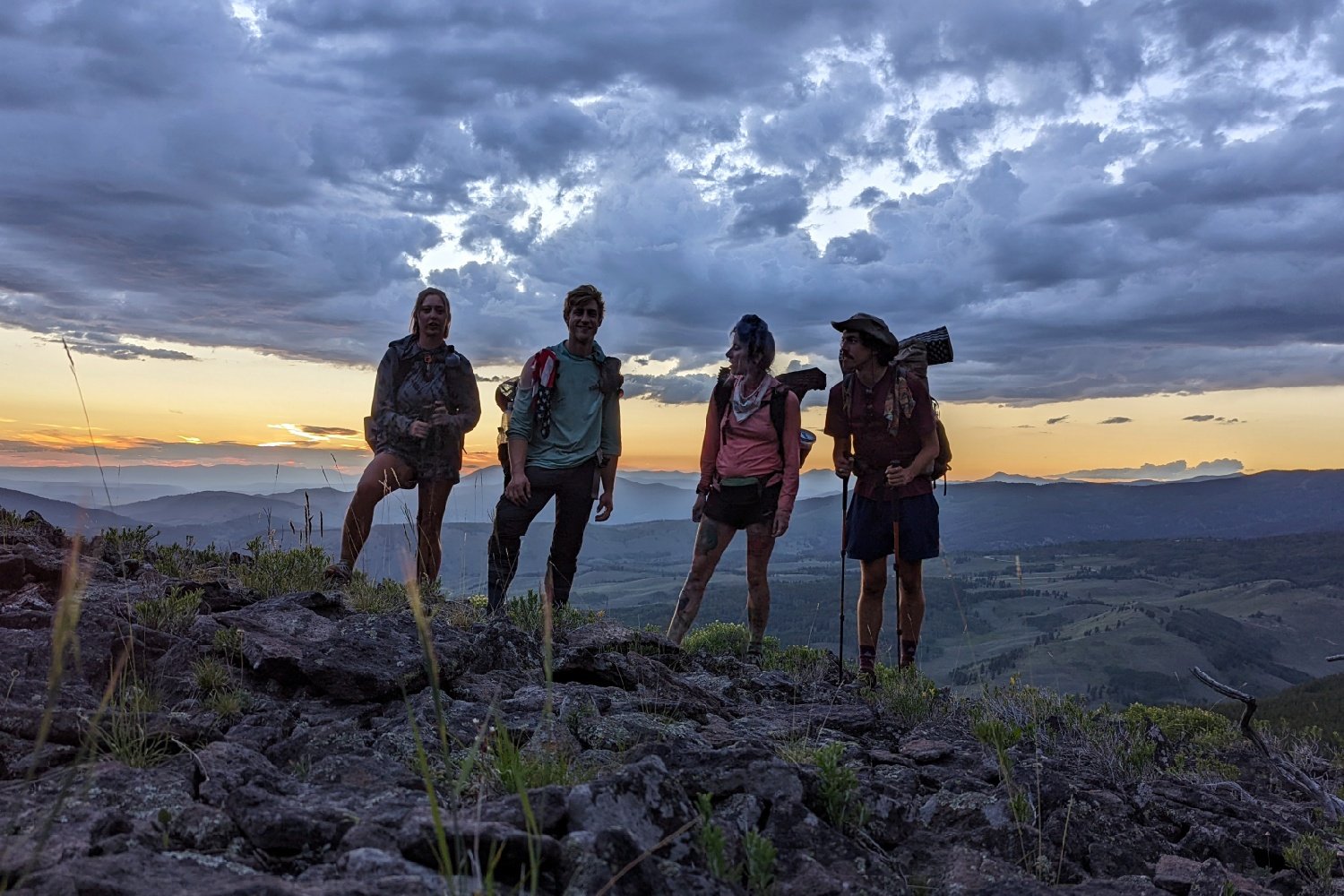 four hikers on a rocky vista with orange sunset and gray clouds overhead.