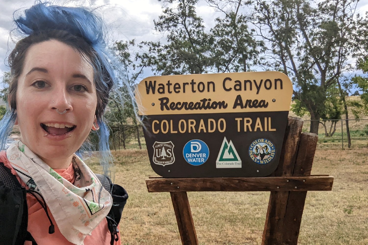 backpacker posing in front of a national forest area sign that says waterton canyon recreation area with the associated managing organizations logos