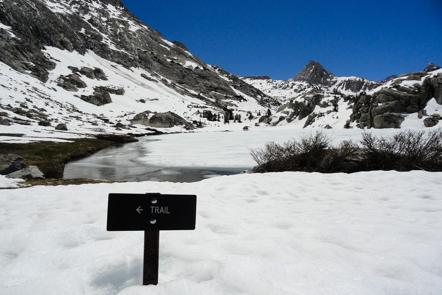 sign pointing to the trail with mountains, snow, and a melting alpine lake behind the sign.