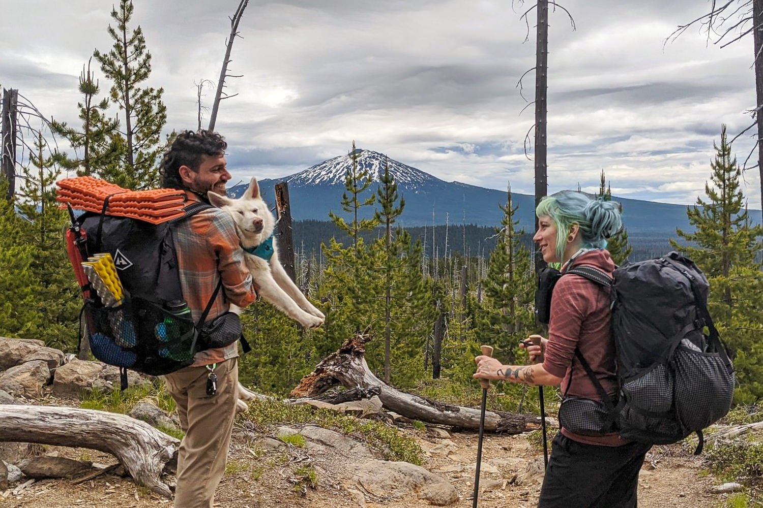 two backpackers with one lifting up their dog in a sweater with mt. bachelor in the distance while on the three sisters backpacking loop