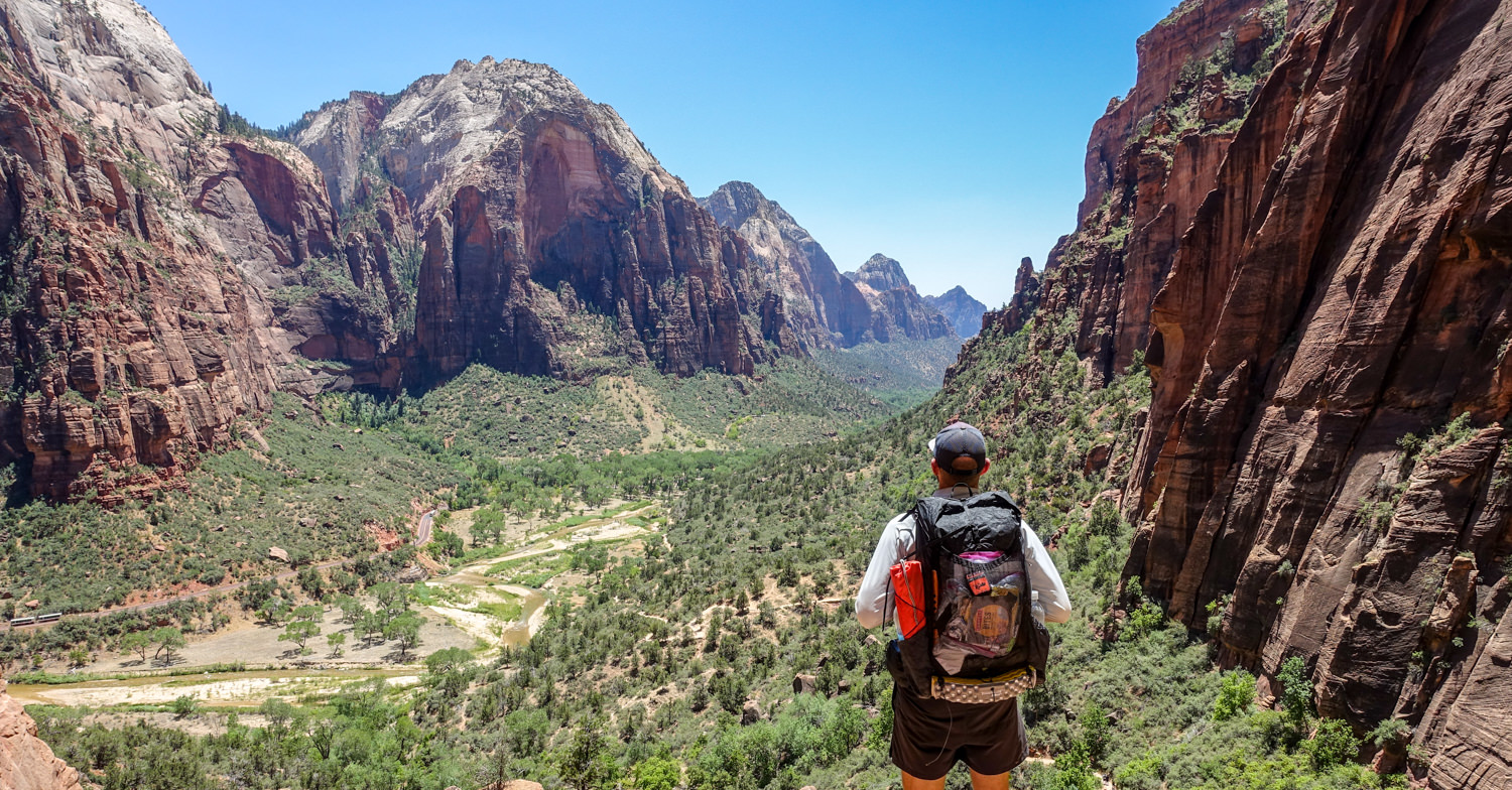 a backpacker in zion national park looks down into zion canyon where zion canyon scenic drive can be seen in the green valley bottom below walls of red and orange canyons walls.