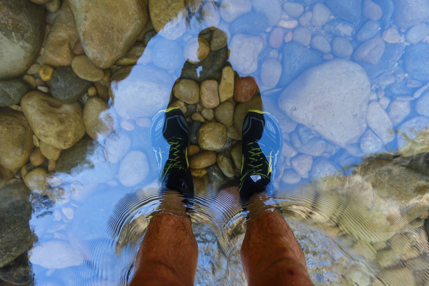 a view of a hikers shoes and socks and feet submerged in the Virgin river on the narrows hike. The water is shallow and clear and you can see the rounded river rock bottom