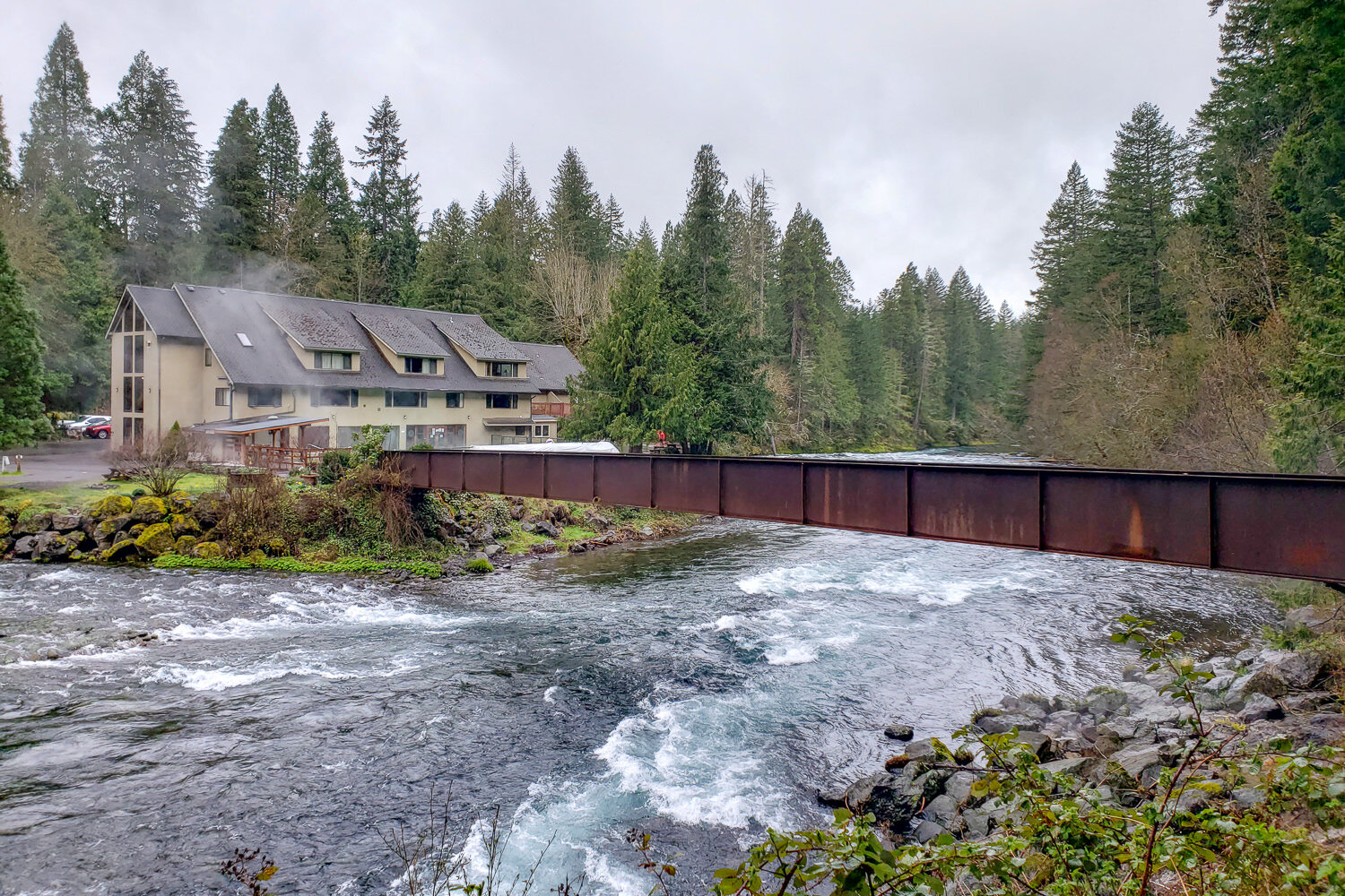 a bridge crossing the mckenzie river leading to the building of belknap hot springs with steam rising from the hot spring pool