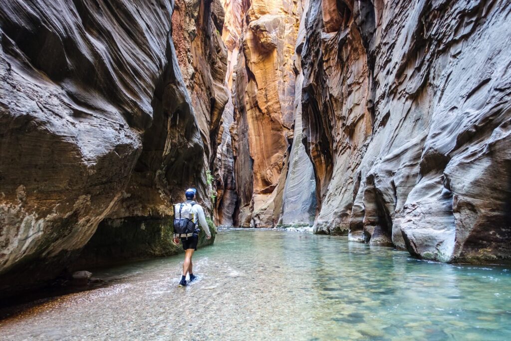 a hiker walking through shallow green-blue water with red sandstone narrows on either side 