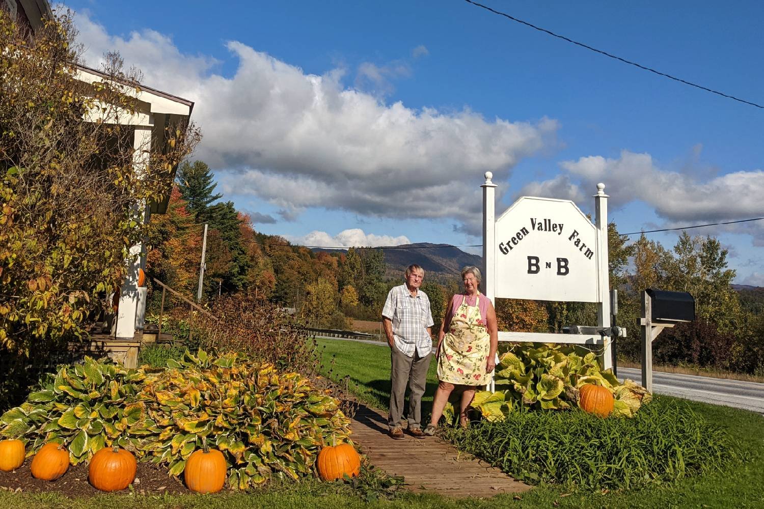 two owners posing in front of their sign that says green valley farm B&B with pumpkins decorating their gardens.