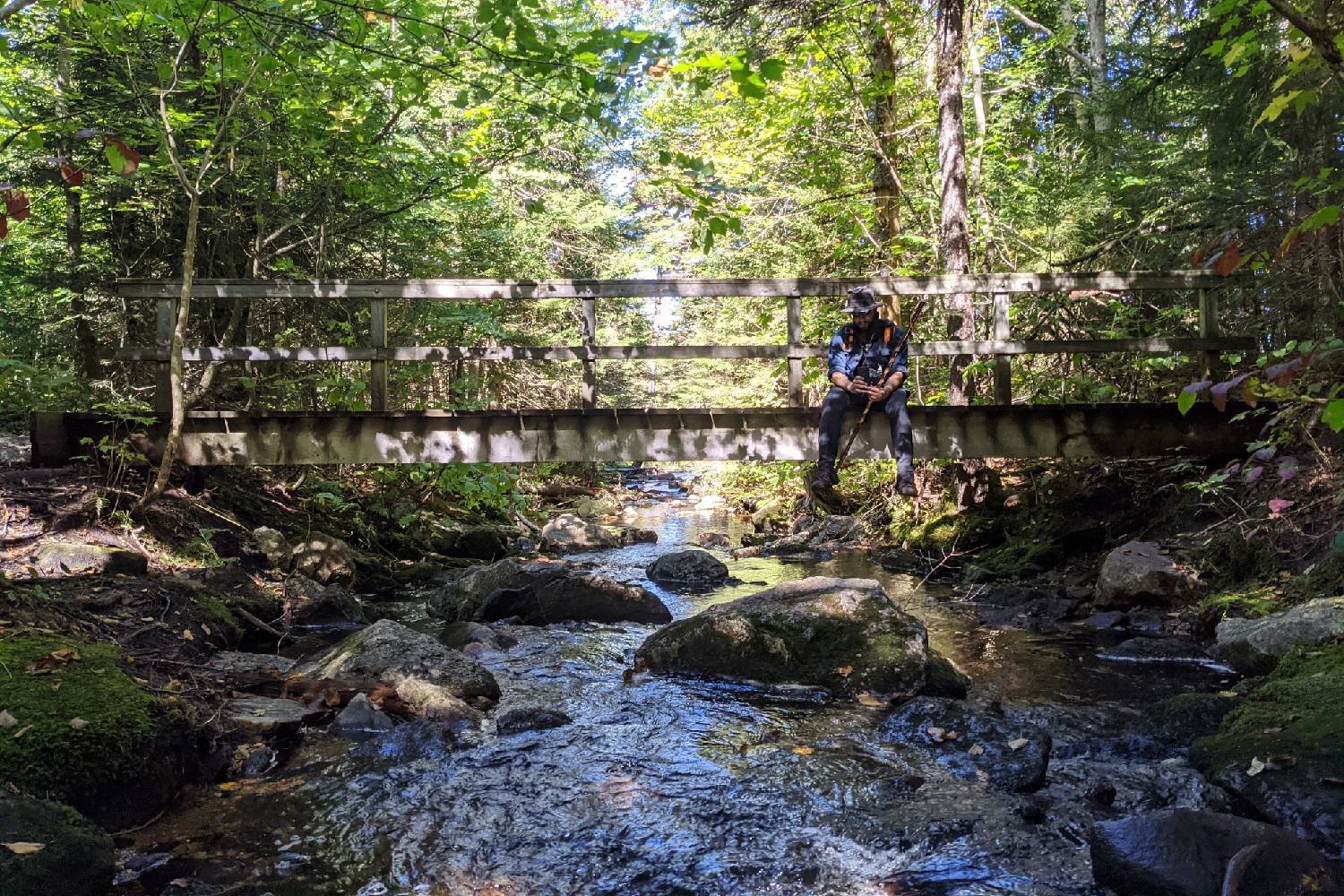 backpacker sitting on a bridge over a river in a green canopy forest.