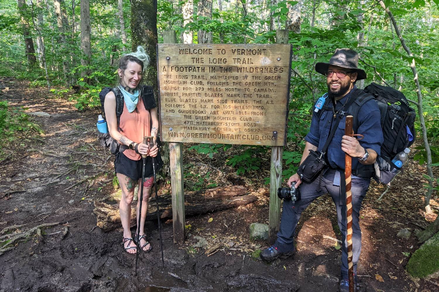 two long trail thru-hikers posing next to an informational sign that says: WELCOME TO VERMONT
THE LONG TRAIL
A FOOTPATH IN THE WILDERNESS
The Long Trail, maintained by the Green Mountain Club, follows the Green Mtn. range for 273 miles from Massachusetts to Canada. White painted blazes mark the trail. Blue blazes mark the side trails. The A.T. follows the L.T. for 105 miles in VT. Maps and guidebooks are available from: The Green Mountain Club,
4711 Waterbury-Stowe Road
Waterbury Center, VT 05677
www.greenmountainclub.org