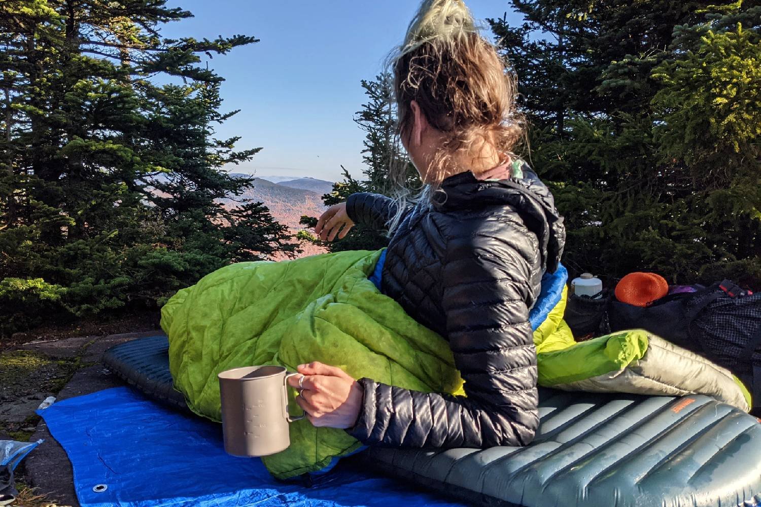 backpacker in the morning holding coffee while laying on her sleeping pad with her backpacking quilt draped over her on a sunny morning