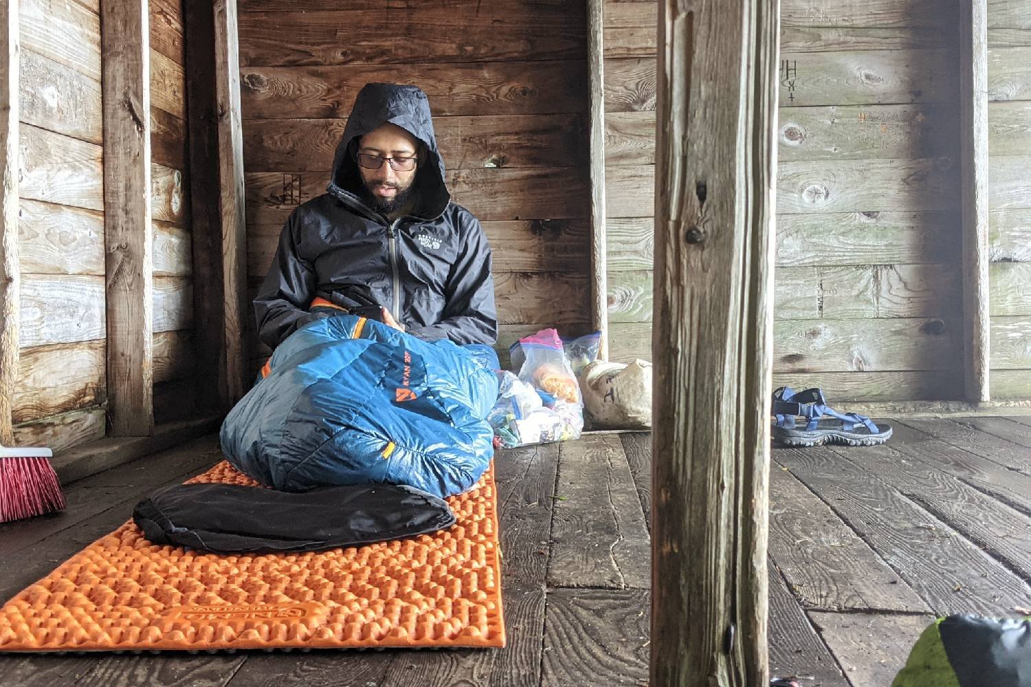 backpacker resting inside a wooden lean-to sitting on his sleeping pad, inside his sleeping bag and wearing his rain jacket.