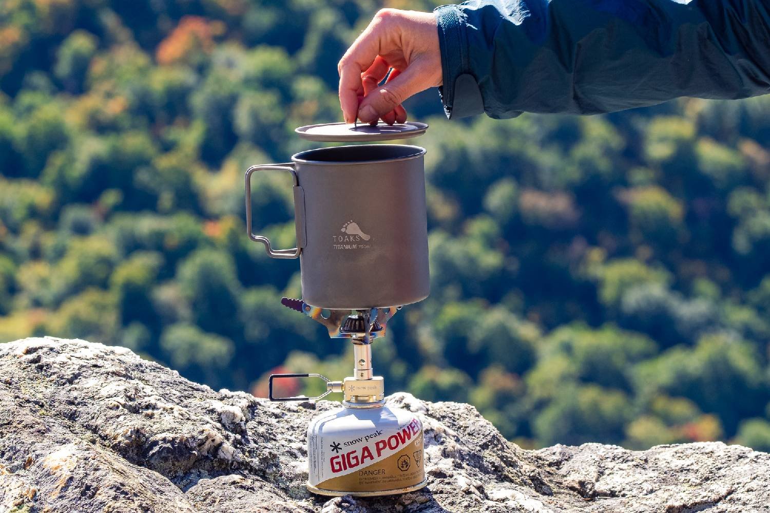 backpacker lifting the lid off their camping stove's pot on a ledge overlooking a tree covered valley