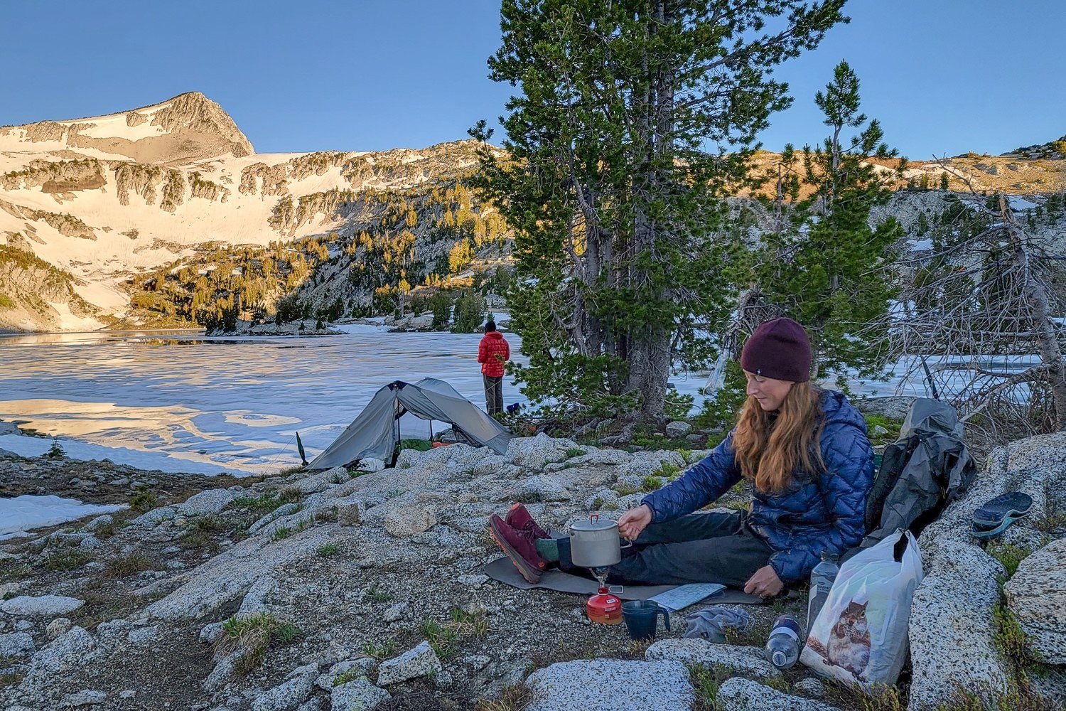 two backpackers in the eagle cap wilderness starting their day with camp coffee and a sunrise view of the snow patched peak above an icy lake. 