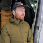 Man with beard wearing a green jacket and baseball cap looks pensively off into the distance while sitting in the open door of a travel van