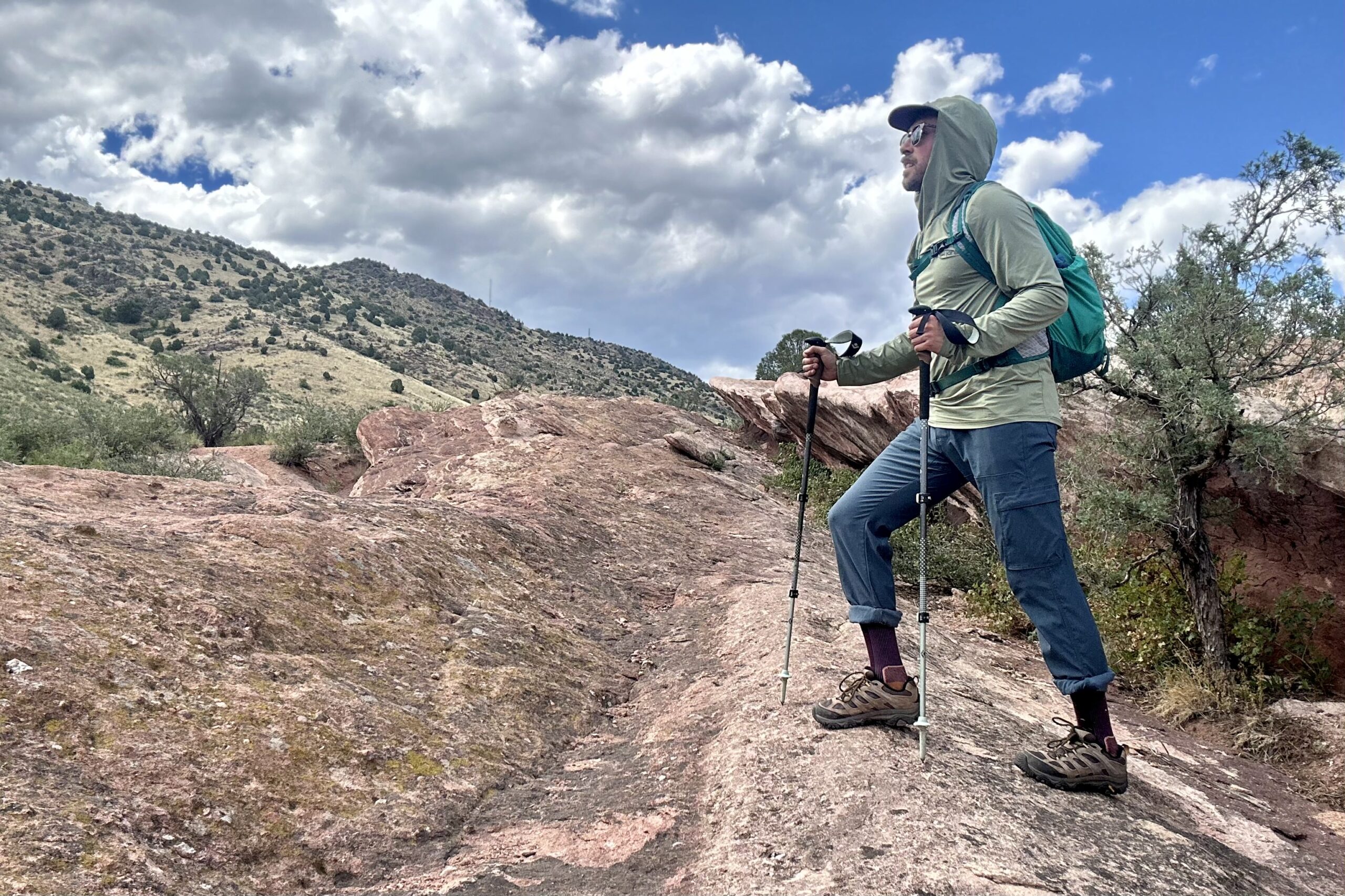 Man wearing a green sun hoody, backpack, pants, and Merrell Moab 3 poses on an uphill hike as he takes in the view of rocks and shrubs.