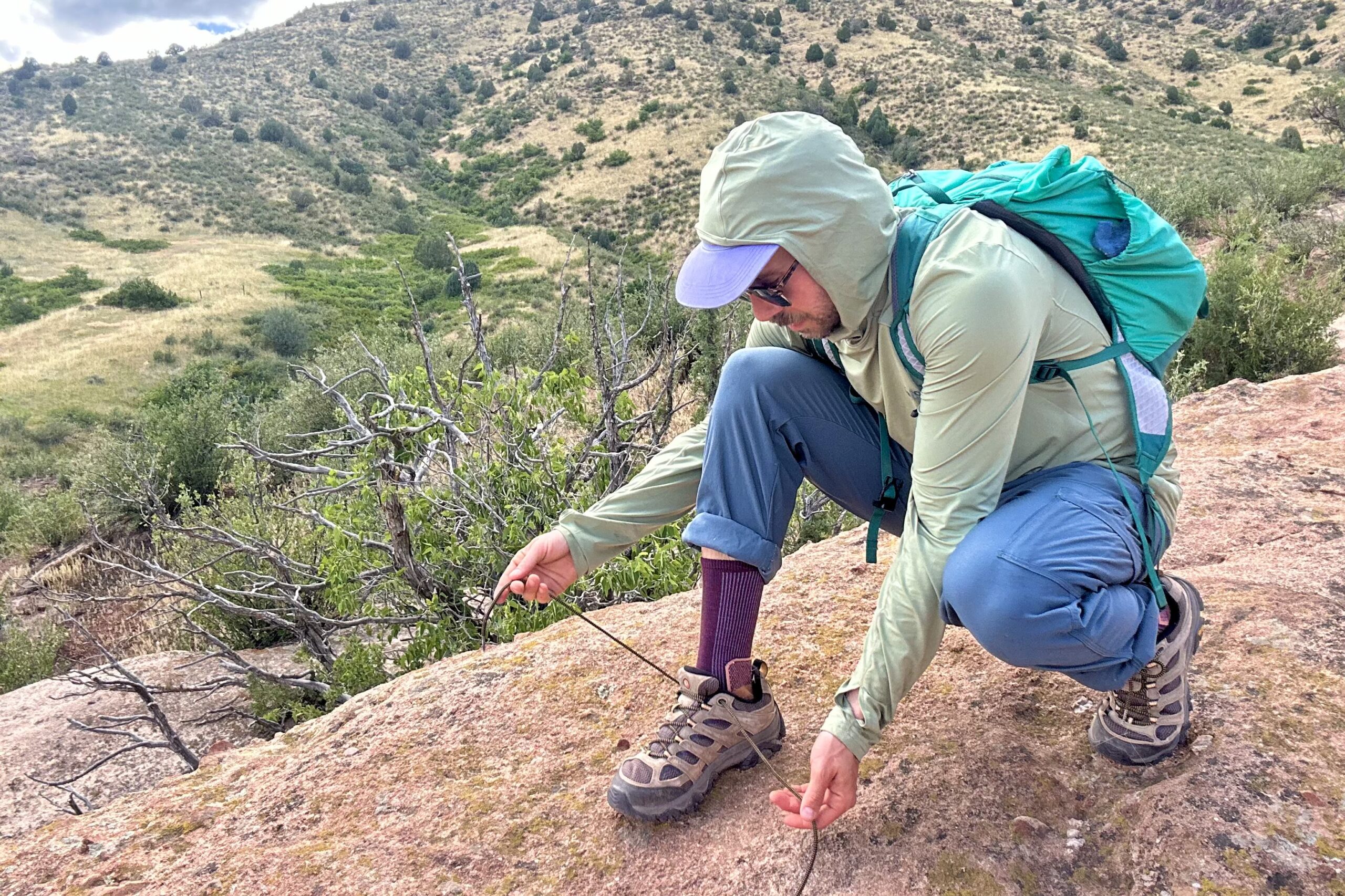 A male hiker ties his shoes in a hunched over position to show off the length of the shoelaces on the Merrell Moab 3s.