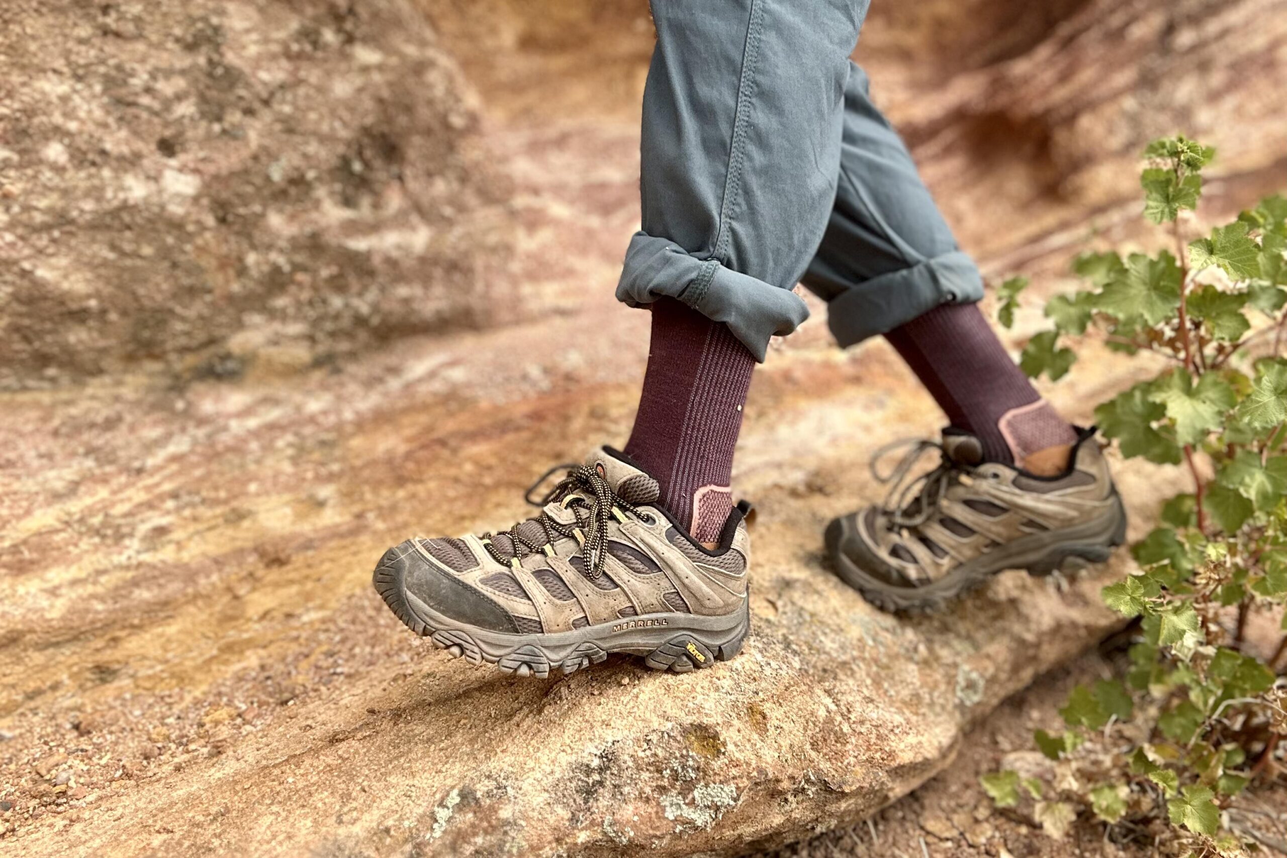 Man's legs from knee down wearing maroon socks and blue pants with Merrell Moab 3 hiking shoes walks along a narrow rocky ledge.