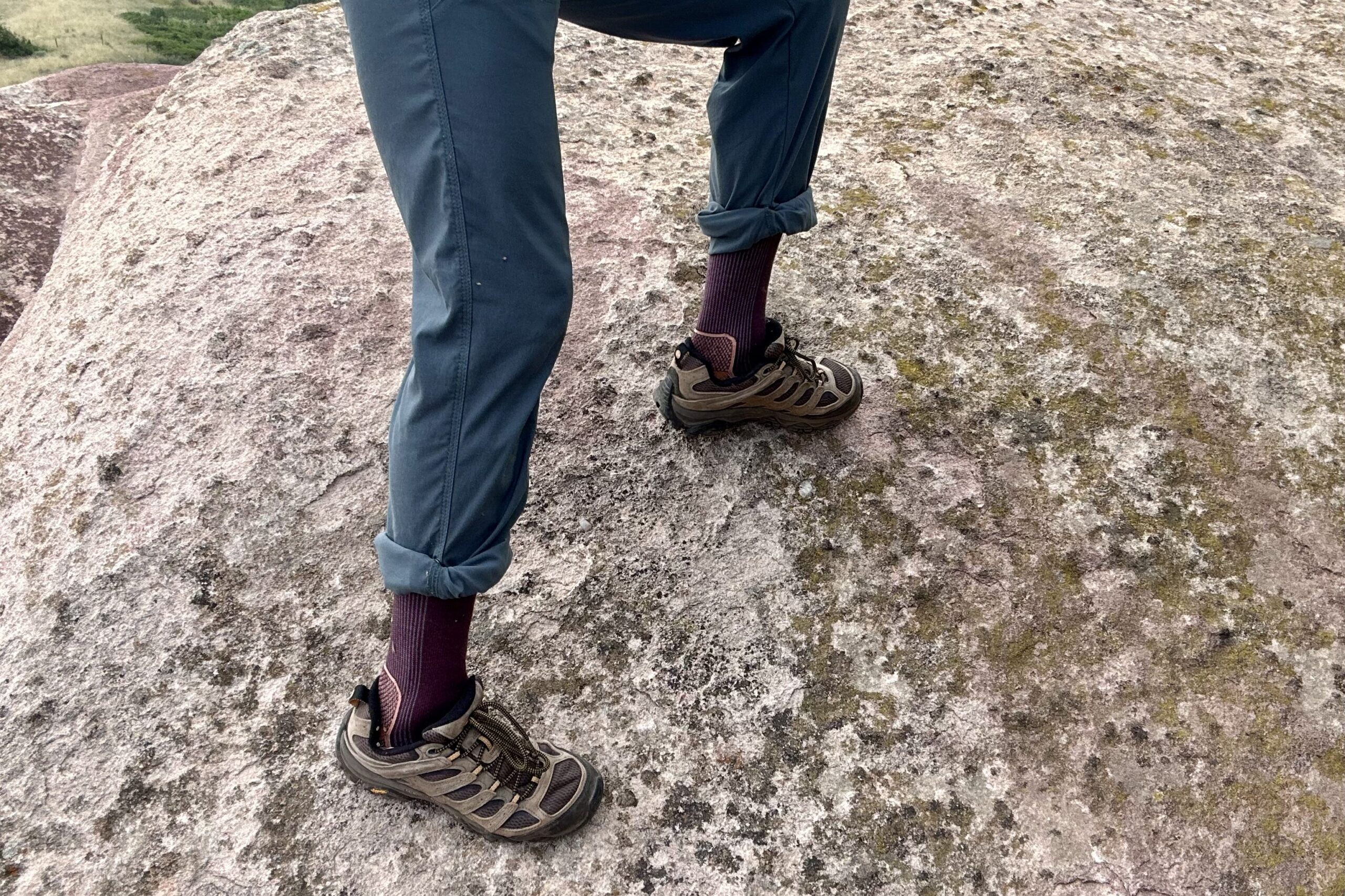 Man climbing a gray rock in Merrell Moab 3 shoes. Shot from the waist from knee down, this man is wearing maroon socks and blue pants with Merrell Moab 3 hiking shoes.