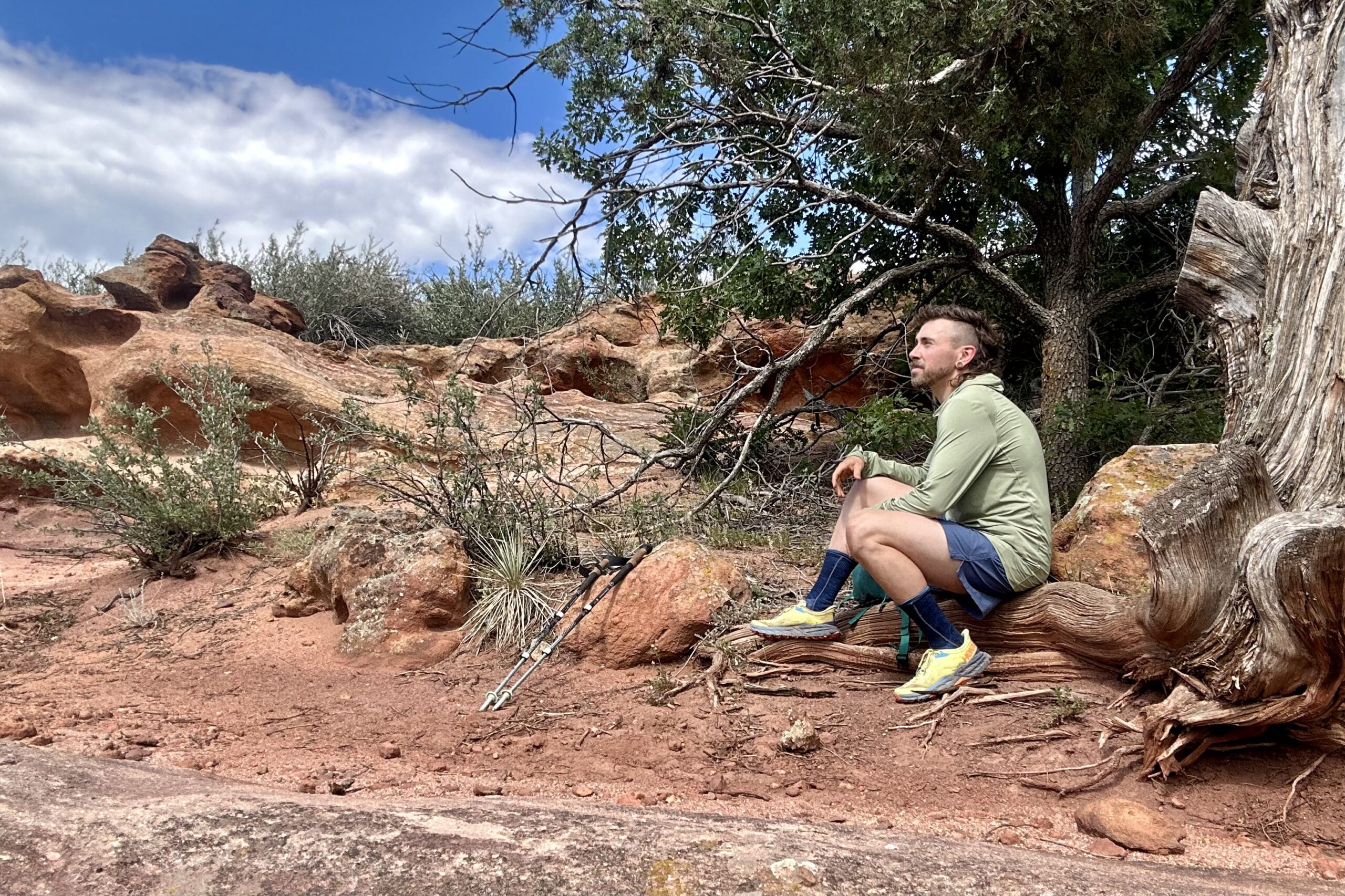 A hiker poses with a backpack, crouching on the ground and taking a break in a desert setting, looking off into the distance.