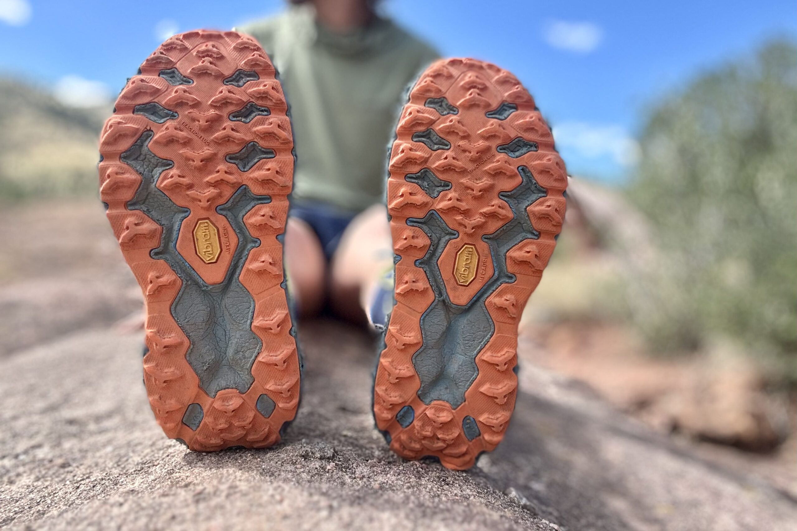 Closeup of trail running shoes looking directly at their outsoles / tread / bottom of shoe, with everything else in background blurred.