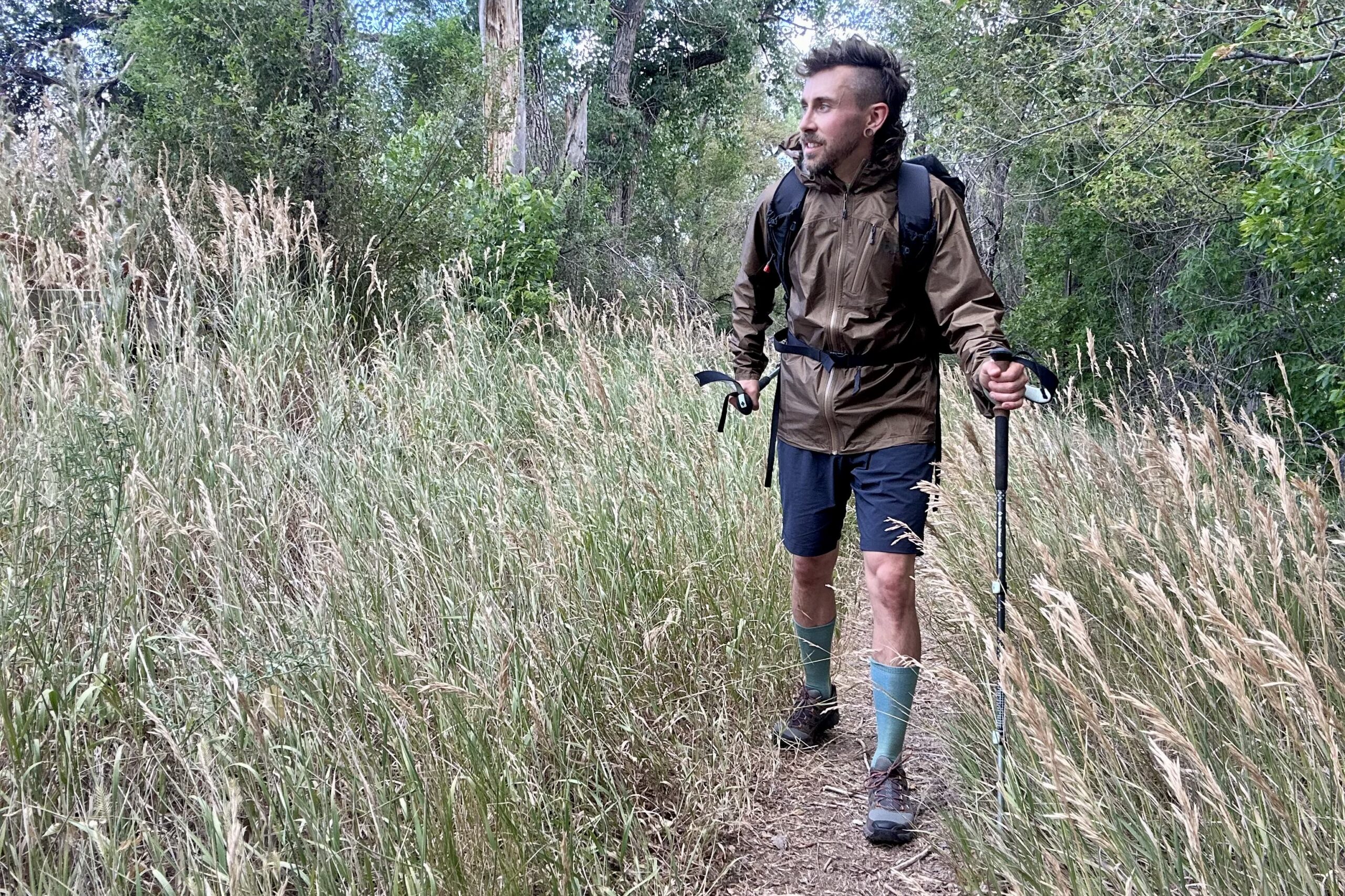 A hiker walks along a trail in a rain jacket and full backpacking gear looking off in the distance in a forest setting.